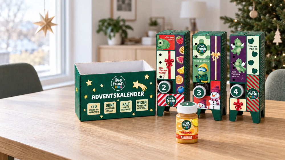 er geöffnete LiveFresh Kids Adventskalender steht auf einem Holztisch vor einem geschmückten Weihnachtsbaum mit der Flasche Orange aus Türchen eins davor. Auf der Verpackung steht "ADVENTSKALENDER" sowie "+20 VITAMINE UND MINERALSTOFFE" "OHNE ZUSATZ VON ZUCKER" "KALT GEPRESST NICHT ERHITZT" und "WISSENSCHAFTLICH ENTWICKELT".