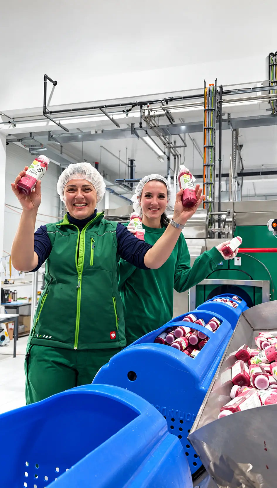 Zwei Mitarbeiterinnen in grüner Arbeitskleidung präsentieren frisch abgefüllte Saftflaschen in der eigenen Manufaktur. Die kaltgepressten Säfte werden mittels HPP Verfahren schonend haltbar gemacht.