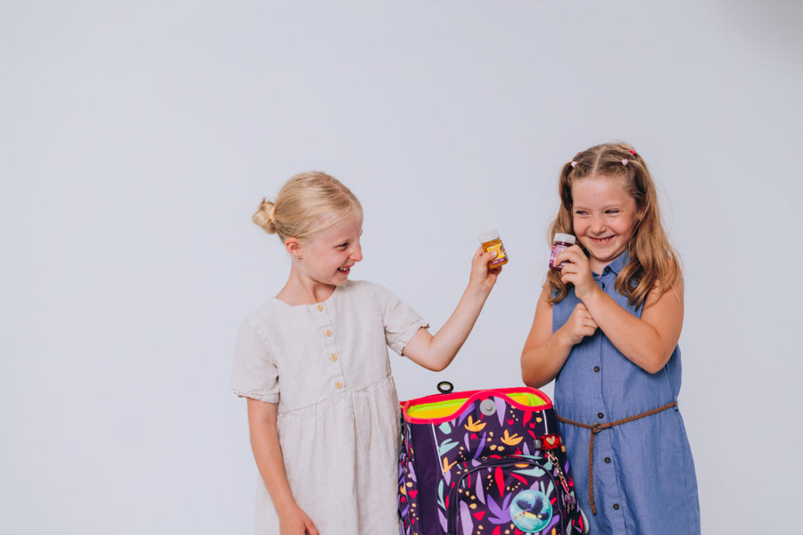 Zwei Mädchen lachen gemeinsam und halten LiveFresh Kids Shots neben einem Schulranzen.