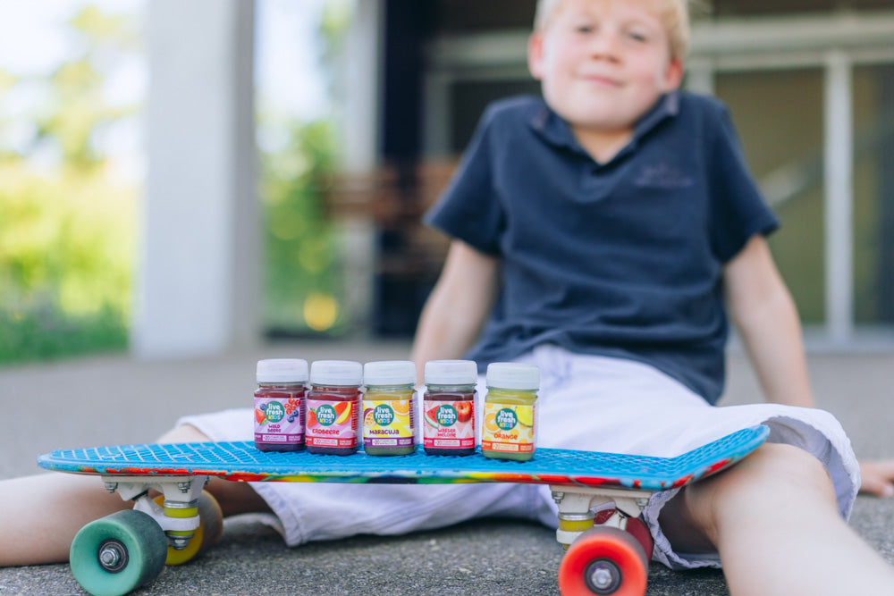 Fünf LiveFresh Kids Shots stehen auf einem Skateboard vor einem sitzenden Jungen.