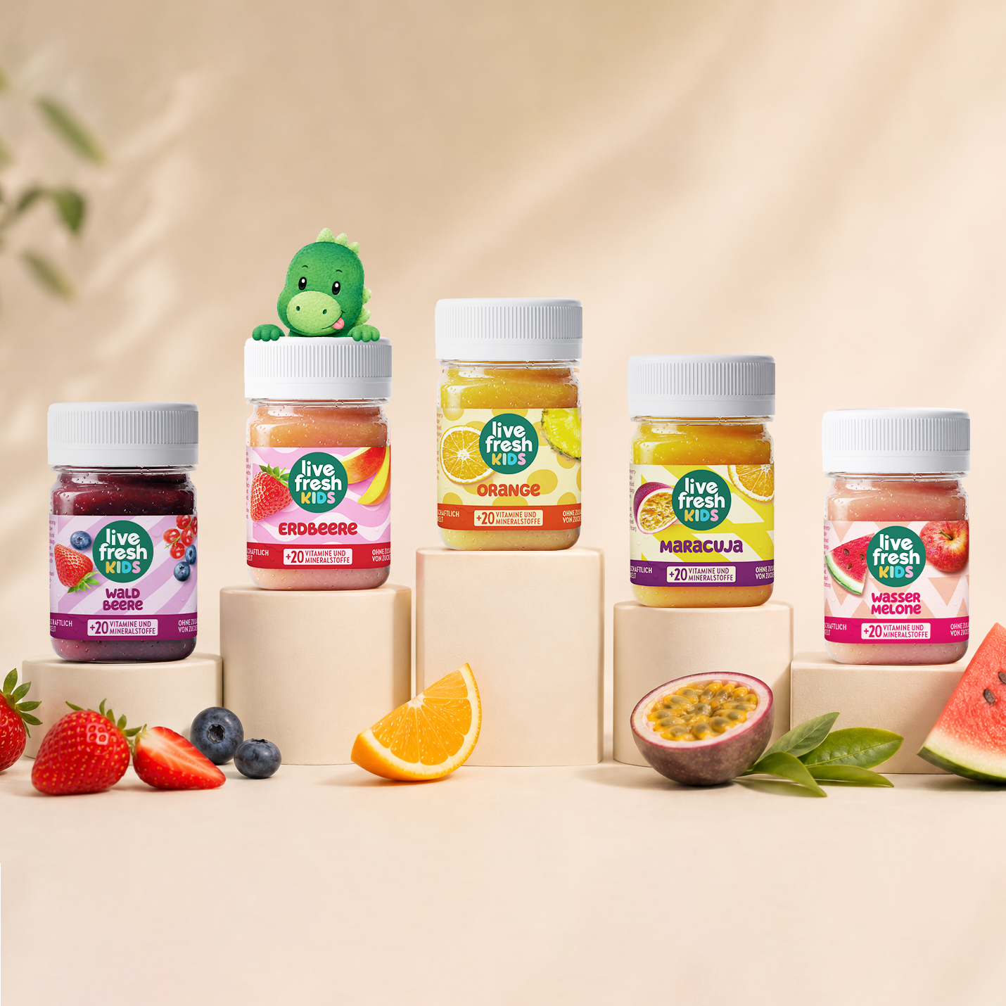 Fünf Gläser mit den Aufschriften "Live Fresh Kids". "Waldbeere". "Erdbeere". "Orange". "Maracuja". "Wassermelone" und "+20 Vitamine und Mineralstoffe" stehen auf unterschiedlich hohen Podesten. Ein kleiner grüner Dinosaurier schaut hinter dem Erdbeer Glas hervor und frische Früchte liegen davor.