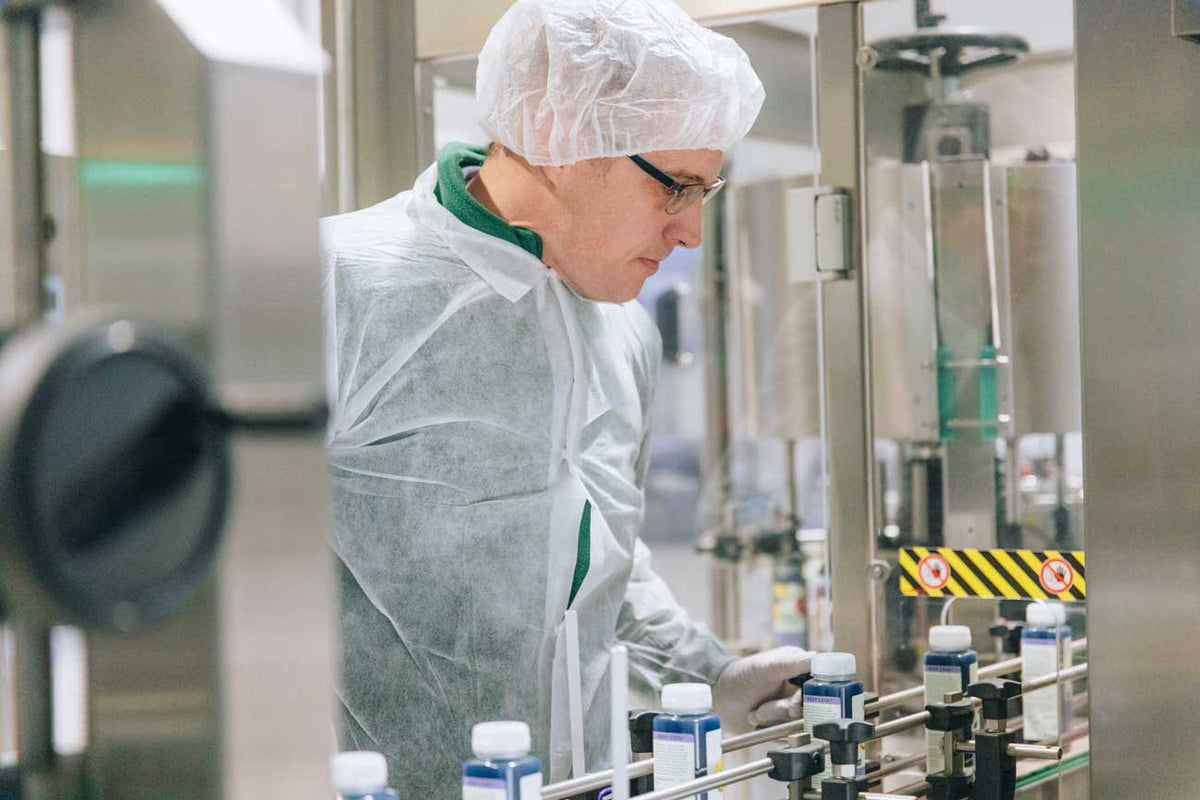 Ein Mitarbeiter mit Schutzkleidung, Haarnetz und Handschuhen überwacht eine industrielle Abfüllanlage. Mehrere kleine Flaschen mit blauer Flüssigkeit laufen auf einem Förderband durch die Produktionsmaschine.